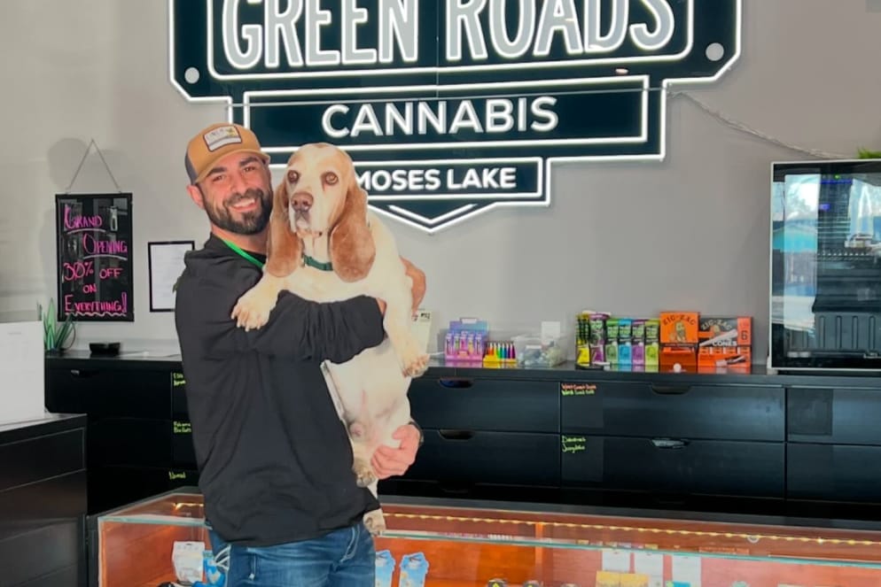 community-3 owner holding basset hound in front of green roads neon sign
